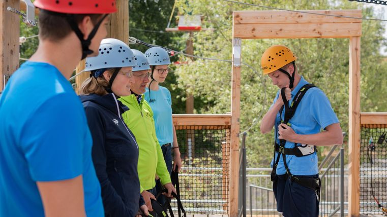 A group of people is standing on a platform and preparing for an adventure activity. Everyone is wearing helmets and safety harnesses.