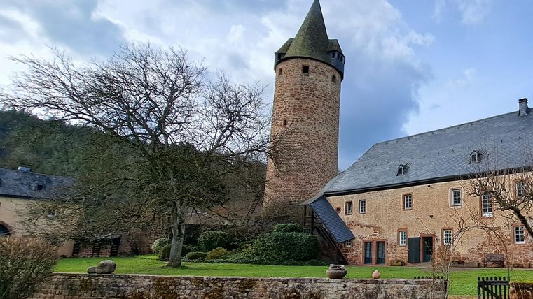 Ein historischer Turm aus Stein steht neben einem alten Gebäude. Der Garten ist grün und gepflegt, umgeben von Bäumen.