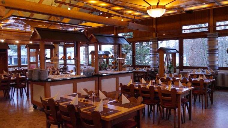 Ein helles Restaurant mit Holzmöbeln und großen Fenstern. Die Tische sind für Gäste gedeckt und die Atmosphäre ist freundlich.