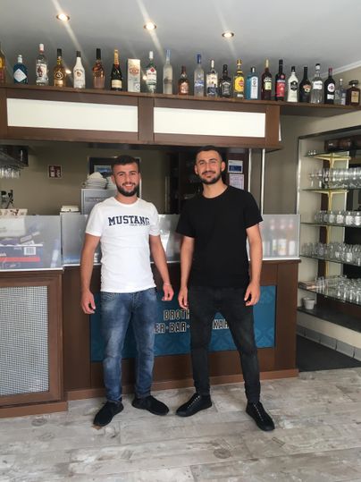 Zwei Männer stehen in einem modernen Restaurant. Im Hintergrund sind Regale mit Flaschen und Getränken zu sehen.