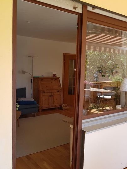 Een blik door de glazen deur naar een lichte woonkamer. Op de achtergrond zijn meubels en een raam met uitzicht naar buiten te zien.