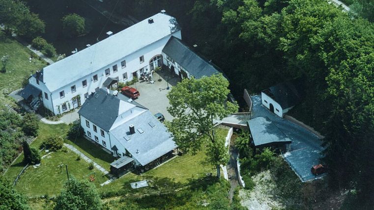 A large building with several extensions, surrounded by trees and green spaces. The courtyard is partially paved and provides space for cars.