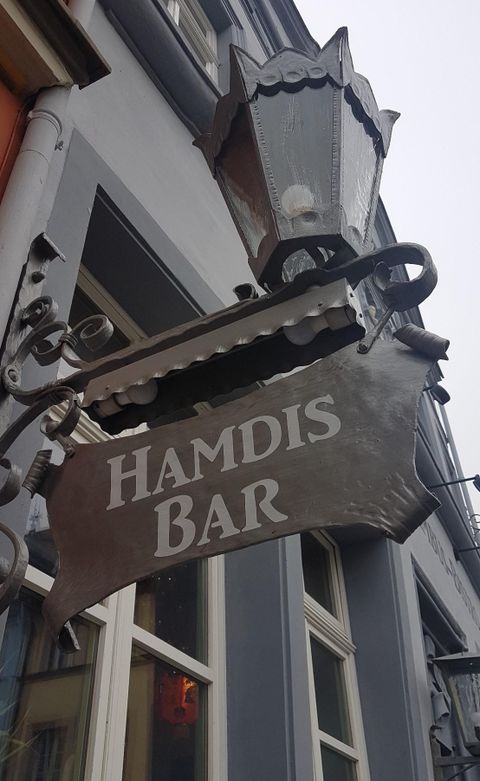 Een decoratief bord van "Hamdis Bar" aan een gebouw. Het bord heeft een stijlvolle vormgeving en is goed herkenbaar.