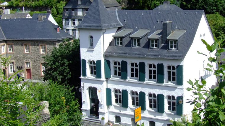A charming, white building with green shutters. Surrounded by trees and other historic buildings.