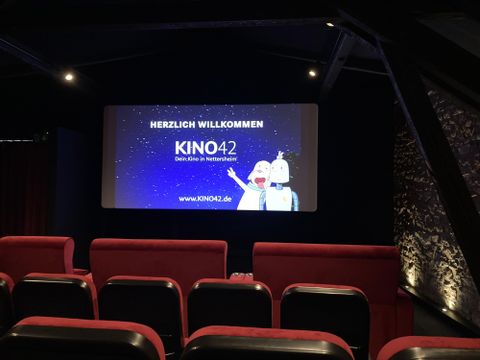 Ein Kinosaal mit roten Sitzen und einer großen Leinwand. Auf der Leinwand steht "HERZLICH WILLKOMMEN KINO42".