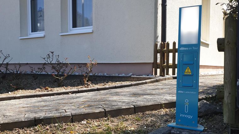 A blue charging station for electric vehicles is on a sidewalk next to a building. In the background, windows and a fence are visible.