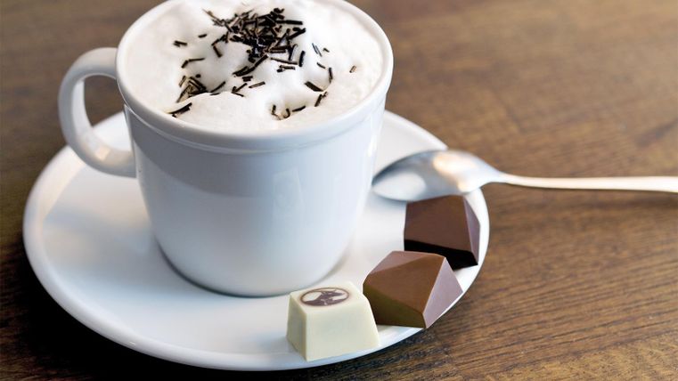 Eine Tasse Kaffee mit Milchschaum und Schokoladenspänen, serviert auf einem weißen Unterteller. Neben der Tasse liegen drei verschiedene Pralinen.