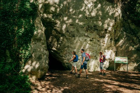 Three hikers enter a cave in a wooded area. The surroundings are green and surrounded by trees.
