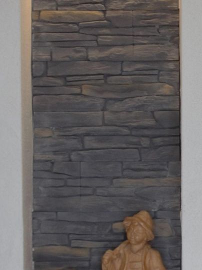 Een muur met een stenen structuur en een houtsnede. Naast staat een houten beeld in een wandelende pose.