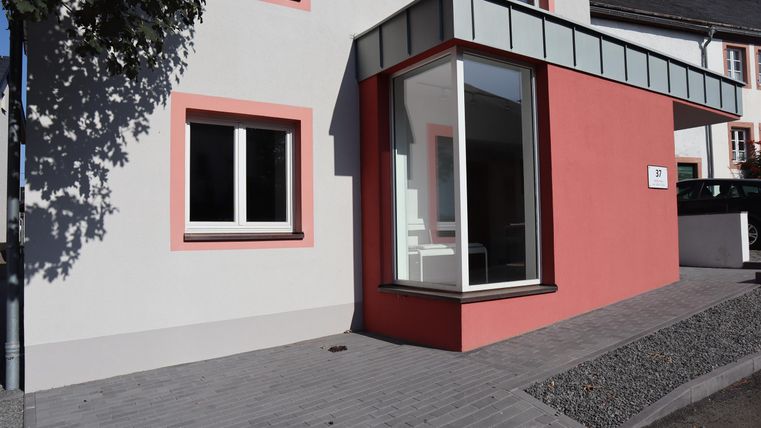 Ein modernes Gebäude mit einer pinken Fassade und großen Fenstern. Der Eingangsbereich ist hell und einladend gestaltet.