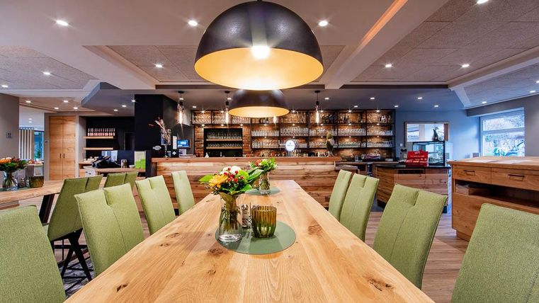 Ein stilvolles Restaurant mit einem langen Holztisch und grünen Stühlen. Die beleuchtete Bar im Hintergrund bietet eine einladende Atmosphäre.