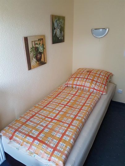Ein einfaches Einzelbett mit einer karierte Decke und einem Kissen. An der Wand hängen zwei Bilder und eine Wandleuchte.