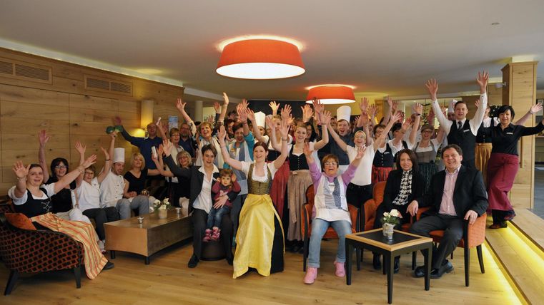 Eine fröhliche Gruppe von Menschen hebt ihre Hände in die Luft. Sie sitzen und stehen in einem modernen Raum mit warmem Licht und holzverkleideten Wänden.
