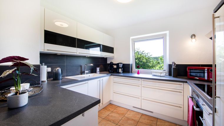 Eine moderne Küche mit weißen Möbeln und einer dunklen Arbeitsplatte. Die Fenster sorgen für viel Tageslicht und Ausblick ins Grüne.