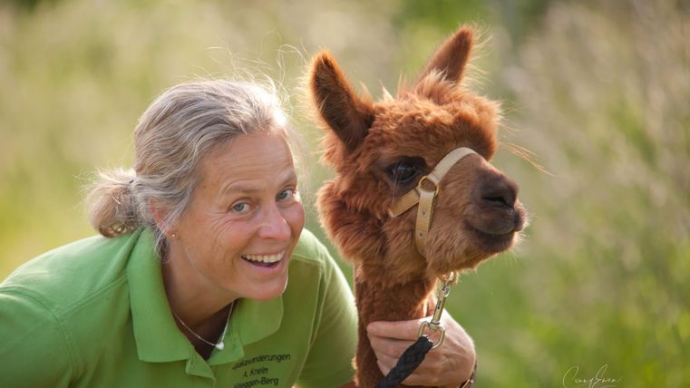 A woman is smiling happily next to a brown alpaca. They are standing in a green environment.