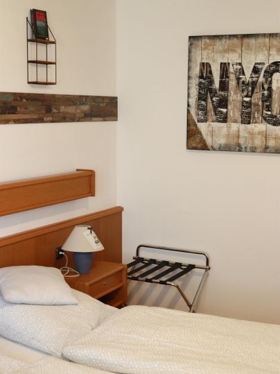 Ein einfach eingerichtetes Zimmer mit einem Bett, Nachttisch und einem Stuhl. An der Wand hängt ein Bild mit der Aufschrift "NYC".