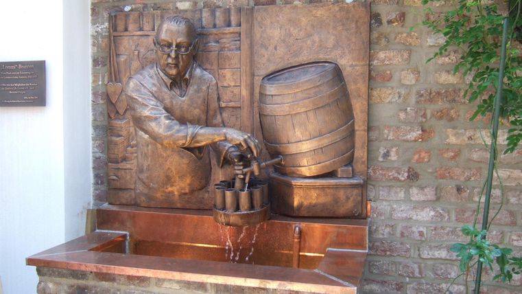 Eine bronze Statue eines Mannes, der mit einer Kelle aus einem Fass schöpft. Im Hintergrund sind Ziegelsteine und Pflanzen sichtbar.