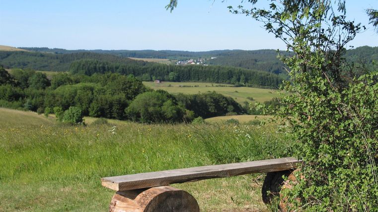 A wooden bench stands on a meadow overlooking green hills and a small village in the background. The surroundings are calm and close to nature.