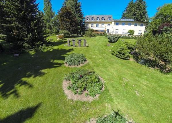 Een uitgestrekte tuin met een goed onderhouden gazon en verschillende planten. Op de achtergrond staat een licht gebouw omringd door bomen.
