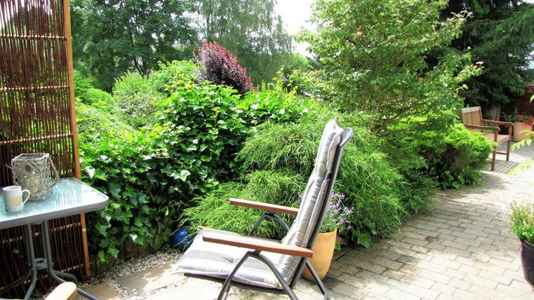 A cozy garden area with a lounge chair and a table. Surrounded by lush greenery and plants.