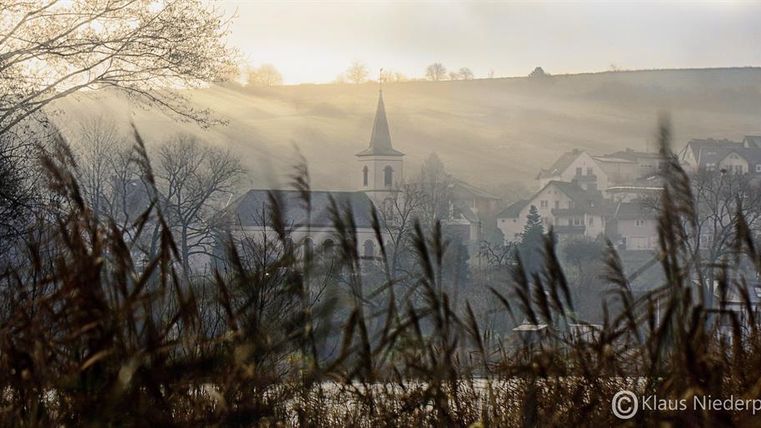 Een schilderachtig landschap met zachte heuvels en een klein dorp in de mist. Voorgrond zijn hoge grassen te zien, terwijl een kerktoren in de verte opvalt.