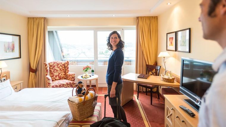 Ein gemütliches Hotelzimmer mit zwei Betten, einem Schreibtisch und einem Balkon. Eine Frau steht lächelnd im Raum und hält ein Gepäckstück.