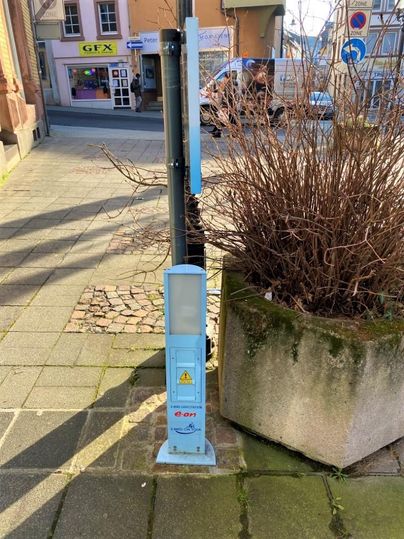 A blue automatic payment unit stands at a street corner. In the background, shops and a traffic light are visible.