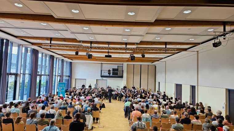 Een grote ruimte met veel zitplaatsen is gevuld met publiek. Op het podium speelt een orkest voor een grote menigte toeschouwers.
