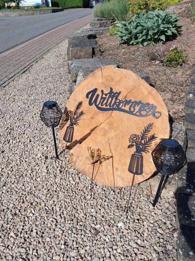 A decorative wooden sign with the inscription "Welcome" stands on gravel. Next to the sign are two black lanterns and some green plants.