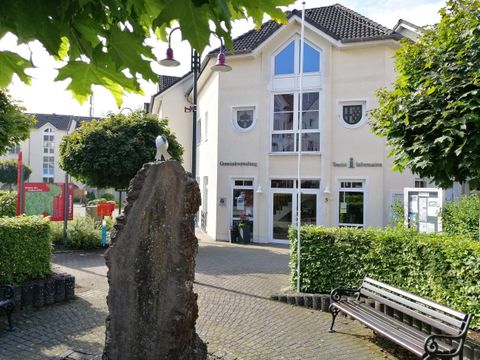 A modern building with large windows and a flag is visible in the background. In the foreground, there are trees and a bench.