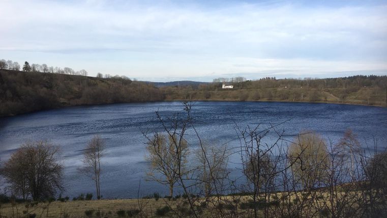 Een rustige meer omringd door zachte heuvels en kale bomen. De lucht is helder en het wateroppervlak weerspiegelt het licht.