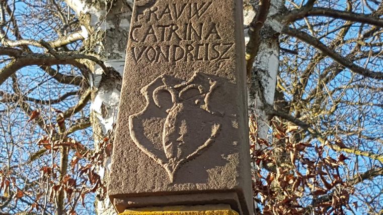 Ein kunstvoller Stein mit Reliefs und Inschriften. Im Hintergrund sind Äste eines Baumes sichtbar.