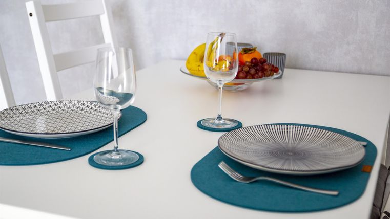 A laid table with two plates, wine glasses, and cutlery. In the background, fresh fruits and a white chair set are visible.