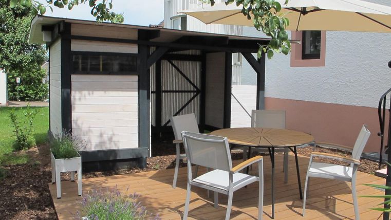 Ein gemütlicher Außenbereich mit einem Holzdeck, Tisch und Stühlen. Im Hintergrund steht ein kleiner Gartenhaus und eine Sonnenschirm schützt vor der Sonne.