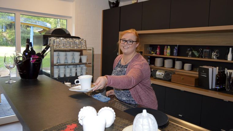 Eine Frau reicht eine Tasse in einer modernen Küche. Im Hintergrund sind verschiedene Geschirrteile und eine Kaffeemaschine zu sehen.