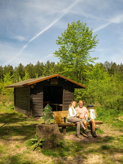 Een stel zit ontspannen op een bank voor een klein houten huisje in het bos. Om hen heen zijn groene bomen en een heldere, zonnige lucht.
