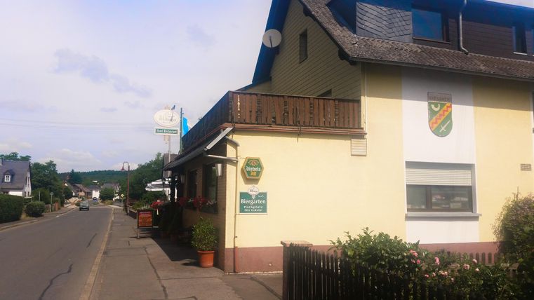 Ein kleines Gasthaus an einer ruhigen Straße. Der Himmel ist leicht bewölkt und es gibt einige Pflanzen vor dem Gebäude.