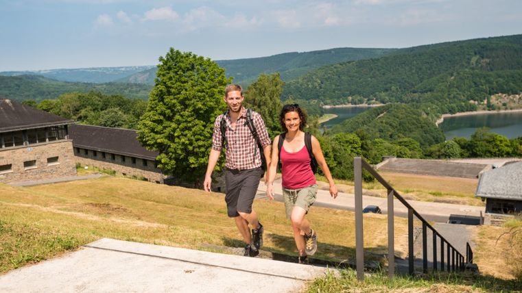 Zwei Wanderer steigen eine Treppe hinauf mit Blick auf Vogelsang IP und die umliegende Landschaft.