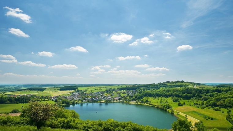 Ein malerischer See umgeben von grünen Wiesen und sanften Hügeln. Blauer Himmel mit vereinzelten Wolken rundet die idyllische Landschaft ab.