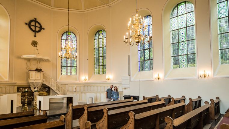 A quiet church with wooden pews and colorful windows. In the foreground, two people are observing the space.