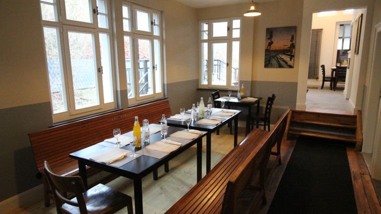 A cozy dining area with long tables and wooden benches. Bright light streams through the windows, creating an inviting atmosphere.