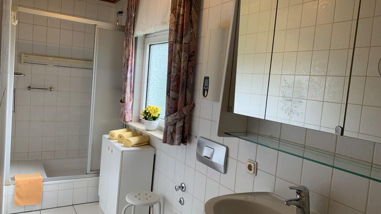 Ein helles Badezimmer mit weißen Fliesen, Dusche und Waschbecken. Es gibt eine kleine Blumendekoration auf der Fensterbank.