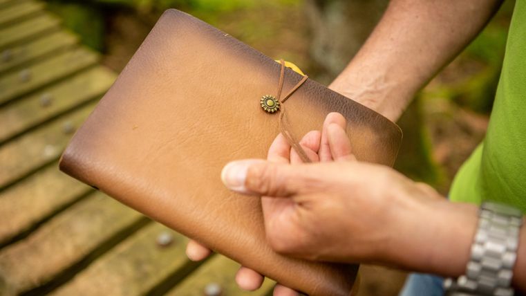 A person is holding a brown leather case with a decorative clasp. In the background, blurred wooden planks and a natural environment can be seen.