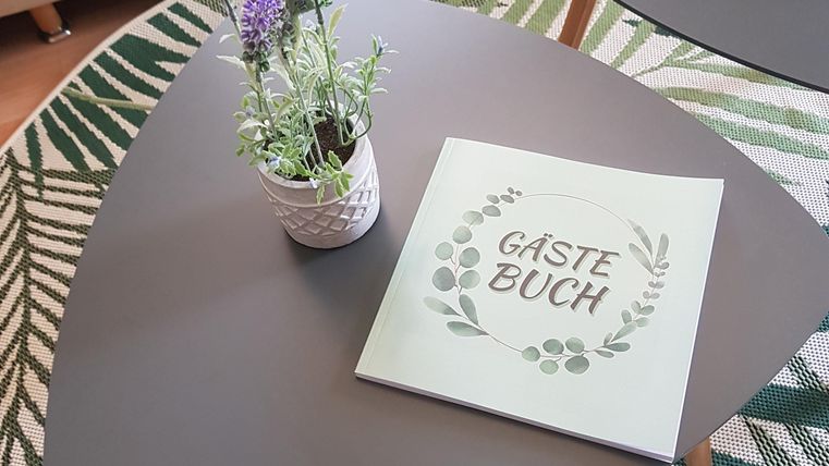 Ein stilvoller Couchtisch mit einem Gästebuch und einer blühenden Pflanze. Im Hintergrund sieht man ein gemütliches Sofa und einen dekorativen Teppich.