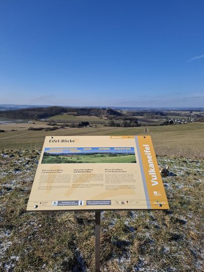 A viewpoint panel stands on a hill overlooking a vast landscape. The sky is clear and blue, and the surroundings are green and gently rolling.