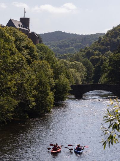 Zwei Kajakfahrer paddeln auf einem ruhigen Fluss. Im Hintergrund sind Bäume und eine alte Brücke zu sehen.