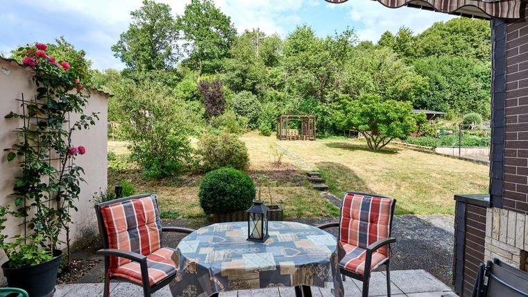 Ein gemütlicher Außenbereich mit einem Tisch und zwei Stühlen. Im Hintergrund ist ein grüner Garten mit Bäumen und Sträuchern zu sehen.