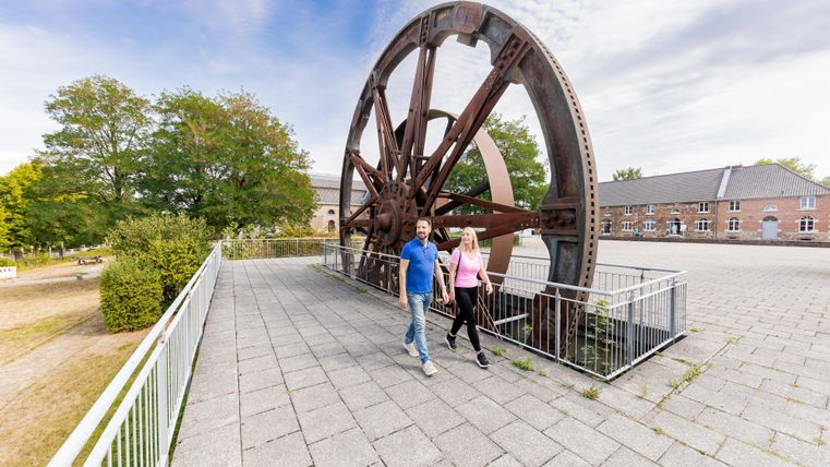 A large historical wheel stands in a park. Two people are leisurely walking on the sidewalk next to the wheel.