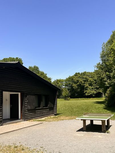 Ein schwarz gestrichenes Holzhaus steht vor einem klaren blauen Himmel. Daneben befindet sich ein Tisch für Tischtennis in einer grünen Wiese.