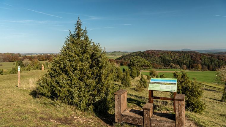 Eine schöne Landschaft mit grünen Hügeln und einem klaren Himmel. Im Vordergrund steht eine Holzbank mit einem Informationsschild.